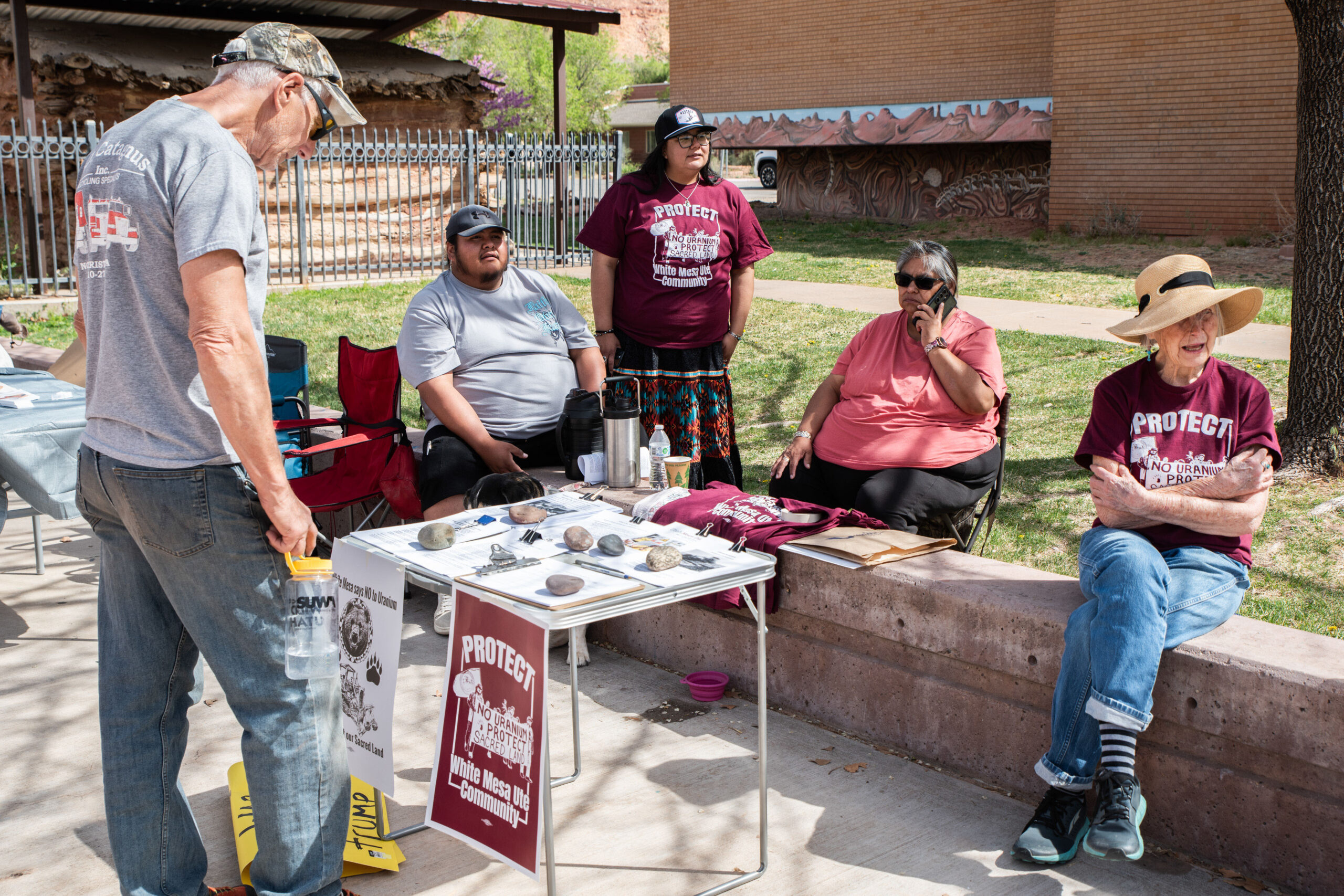 A group of people sit near an information booth set up at a rally. Several are wearing red shirts with white text that read, "Protect White Mesa Ute Community."