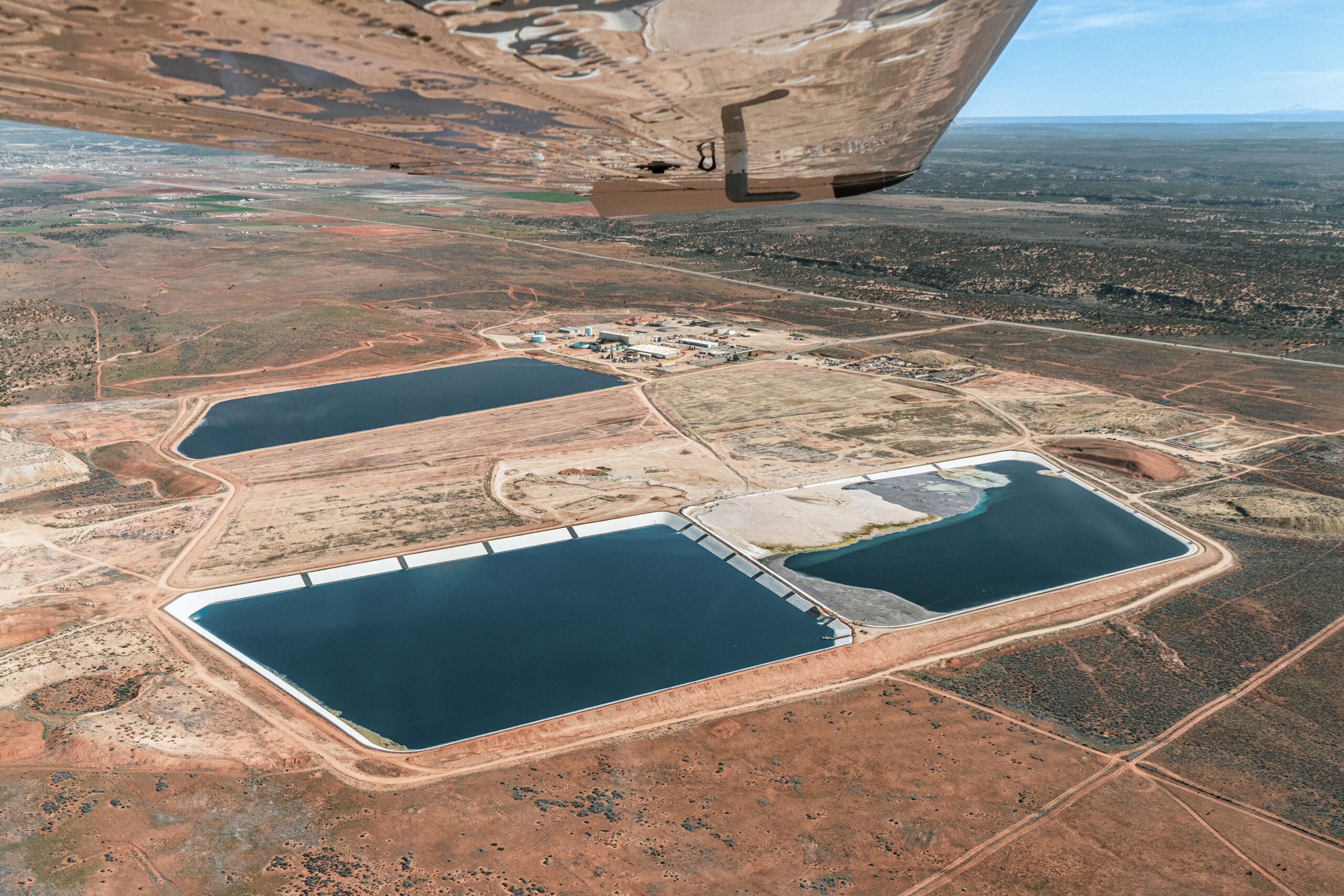 An aerial view shows tailing ponds amidst a mesa landscape. The wing of an airplane from which the photo was taken is seen in the frame.
