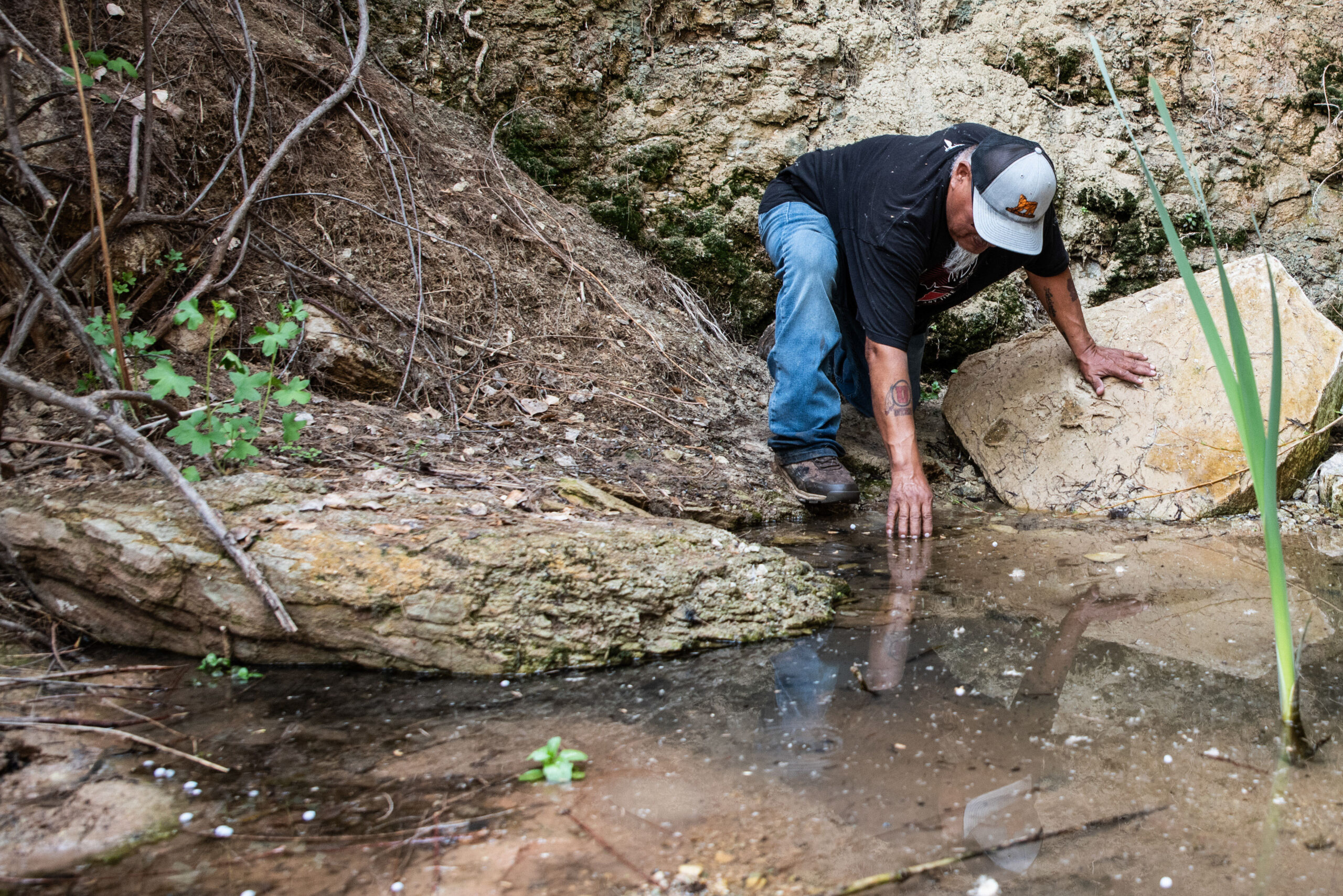 A photo of Malcolm Lehi reaching down to touch the water of Entrance Spring.